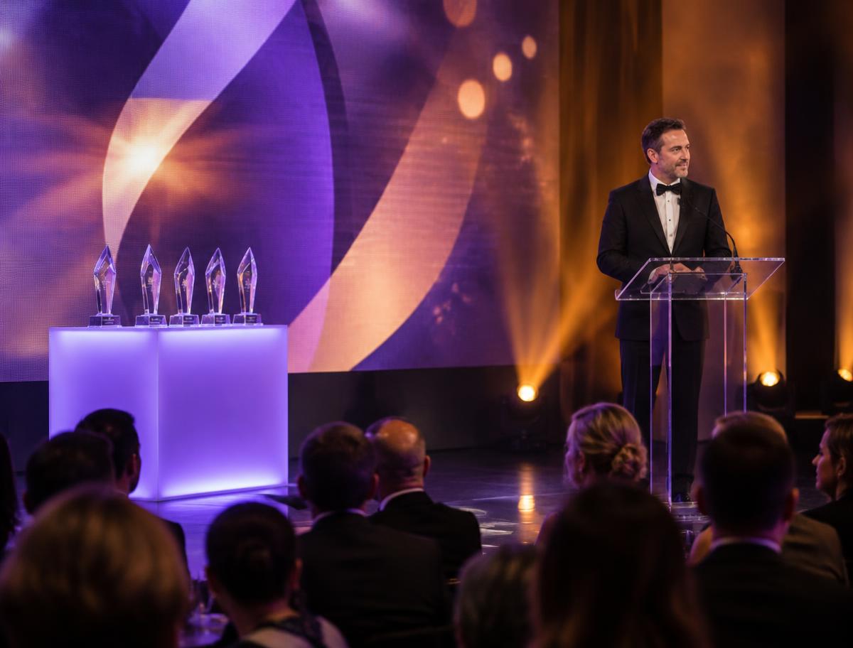 Presenter at a glass lectern beside illuminated trophies on stage